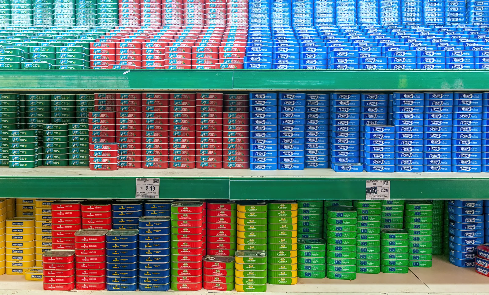 Lineal de un supermercado con latas de sardinas de varias marcas apiladas por colores, perfiles portaprecios verdes y etiquetas de precio